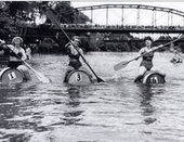 Old photo of water carnival at Healdsburg Veterans Memorial Beach with bridge in background and 9 women rowing barrels-170