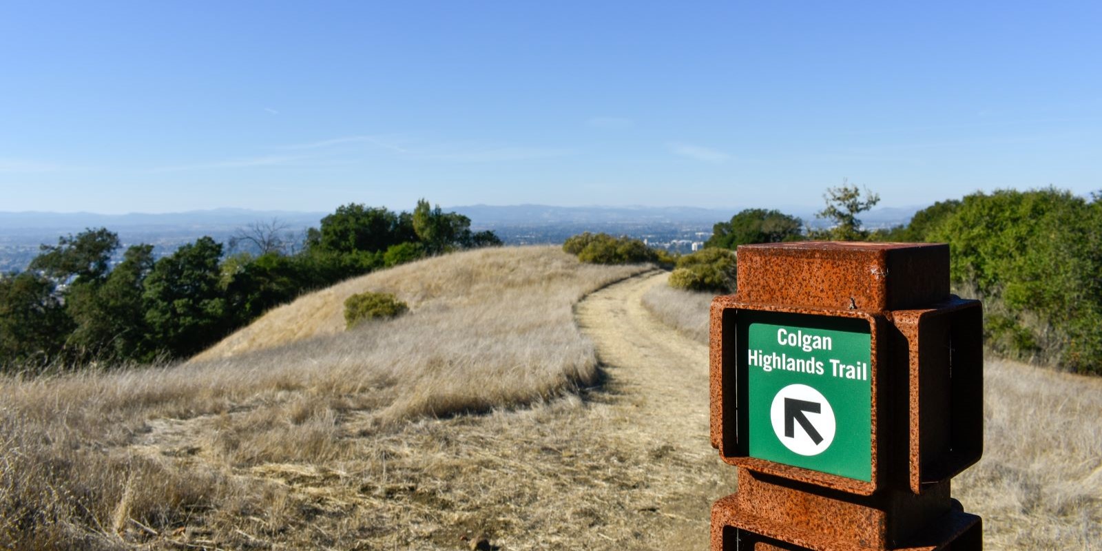 Trail sign for Colgan Highlands Trail in Taylor Mountain Regional Park, with a dirt path curving through dry grassland and panoramic views of Santa Rosa and distant hills in the background