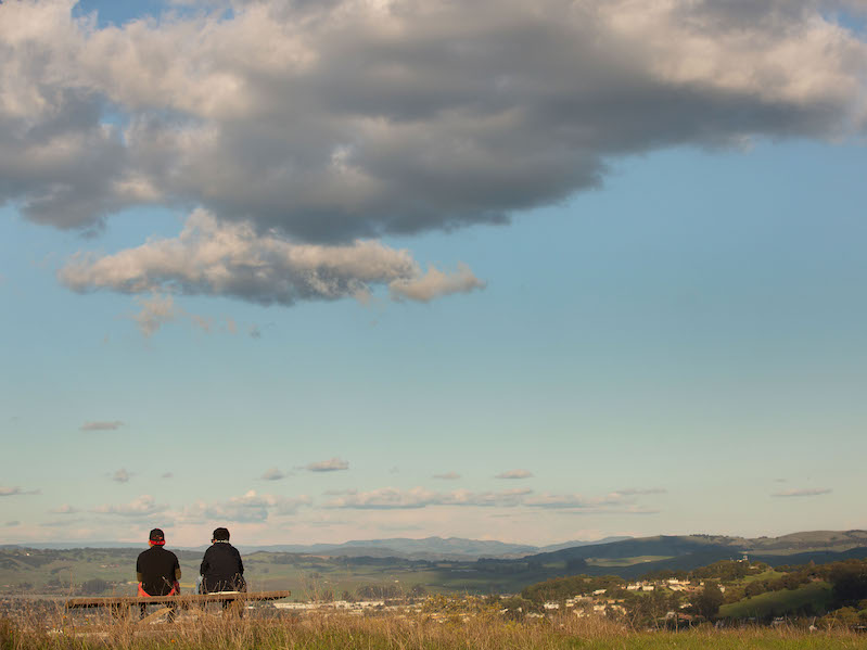 Two people sit on a bench with view of sky and rolling hills